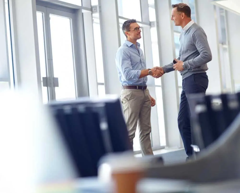 Zwei Männer im Business-Outfit begrüßen sich mit einem Handschlag in einem modernen Bürogebäude – ein typisches Motiv für berufliche Zusammenarbeit und Networking beim Arbeiten in der Schweiz.