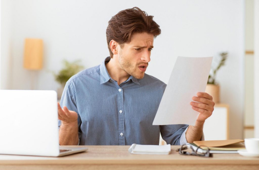 Mann sitzt fassungslos vor seinem Laptop und hält ein Dokument in der Hand – zeigt Unsicherheit über Ansprüche oder Leistungen der Arbeitslosenversicherung Schweiz.