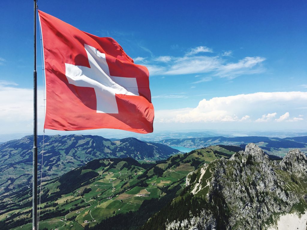 Schweizer Flagge weht auf einem Gipfel in den Alpen, mit Blick auf grüne Täler und einen See.