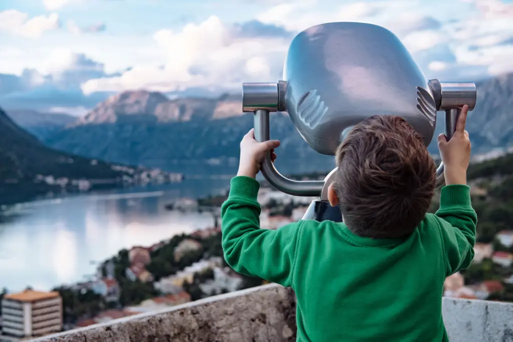 ein kleiner Junge in grünem Pullover blickt durch ein großes Fernrohr auf eine schweizer Stadtlandschaft mit Bergen und Wasser.