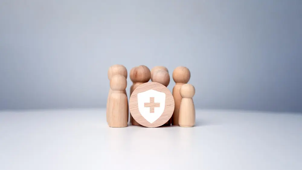 Holzfiguren symbolisieren eine Familie, umgeben ein Schild mit Kreuz – sinnbildlich für Sicherheit durch einen guten Krankenversicherungsvergleich.