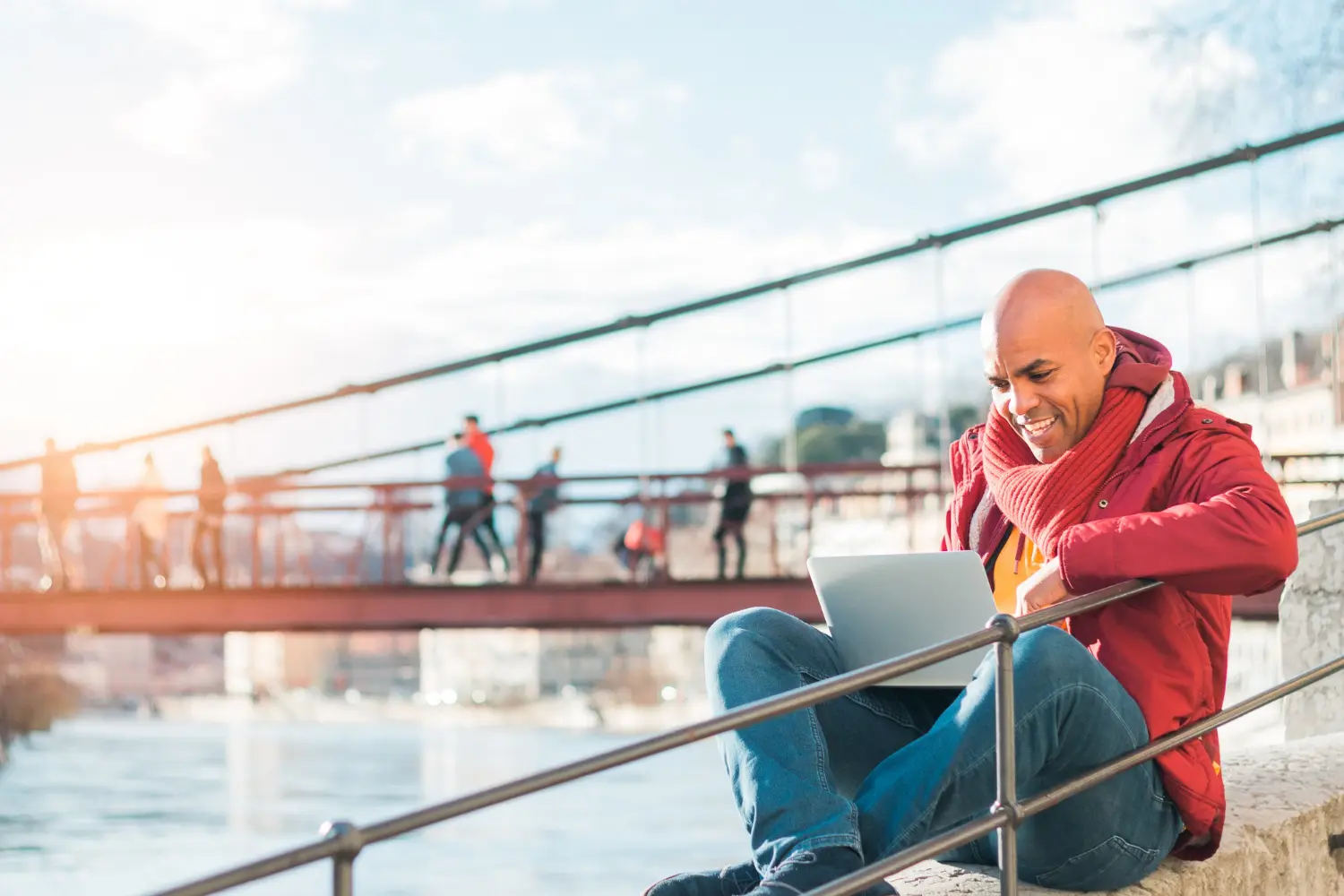 Lächelnder Mann arbeitet entspannt mit dem Laptop im Freien vor einer Brücke – symbolisch für moderne und flexible Arbeitsbedingungen in der Schweiz.